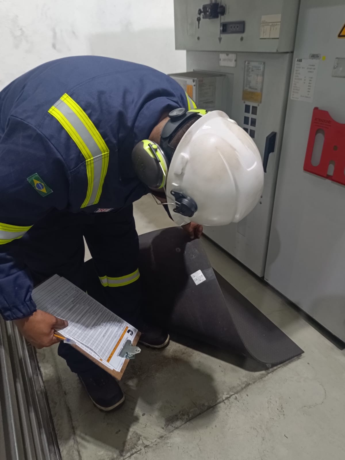 Profissional com uniforme azul, capacete branco e prancheta realizando uma inspeção técnica.