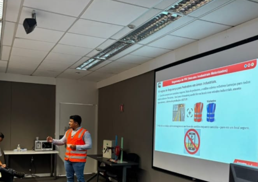 Instrutor com colete laranja conduzindo um treinamento presencial junto à apresentação projetada.